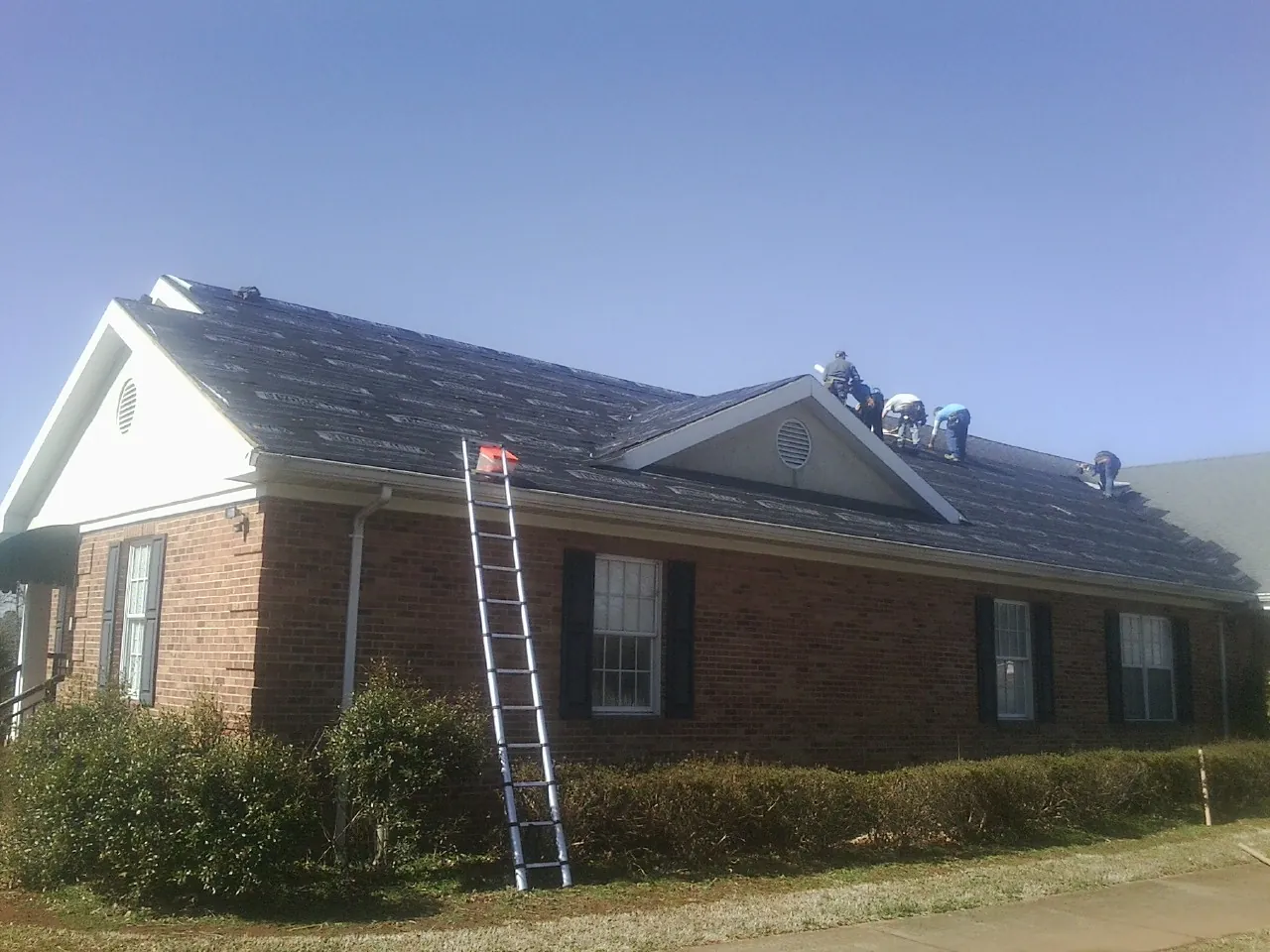 Crew tearing shingles off a brick ranch roof with a ladder set — Emergency Roof Repair work by our crew in Aplington