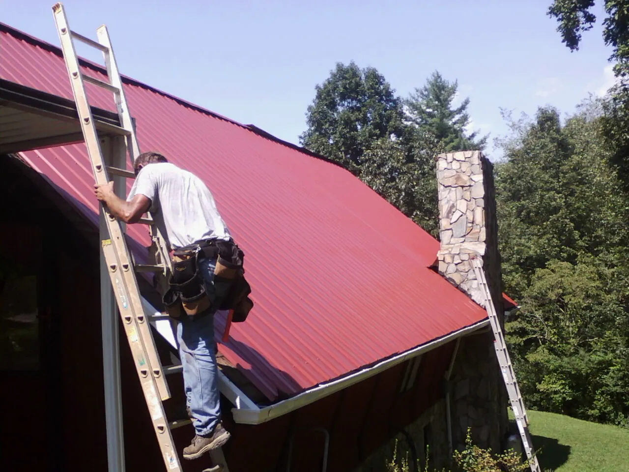 Roofer on a ladder beside a red metal roof and stone chimney — Soffit & Fascia Repair by Roofing Brothers in Aplington